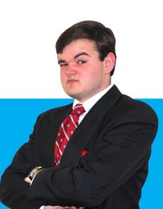 Picture from the torso up of a man wearing a business suit with a background of a solid light blue stripe and white above. The man is wearing a watch on his left wrist and a red tie and pocket square. He bears a quizzical expression with one eyebrow raised.